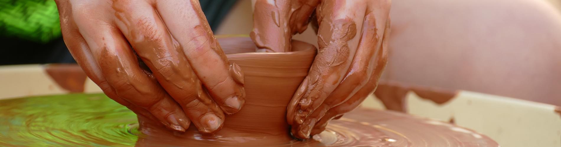 Young person sits over a pottery wheel, crafting a small pot with wet clay.