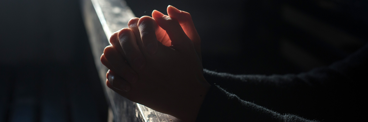 Two hands are grasped together, in shadow, leaning on a church pew