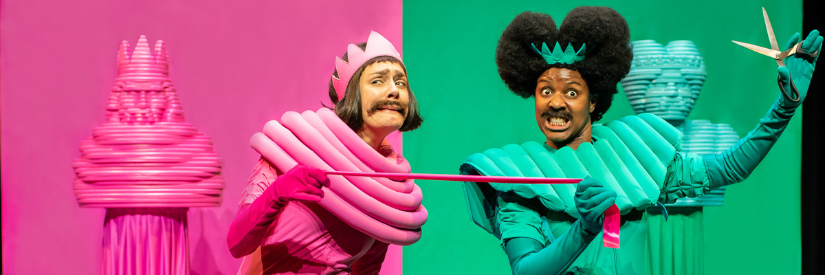 A wacky image of two people with, large collars, long gloves and crowns, one wears all bright pink and one bright green. They both hold a piece of pink fabric, the person dressed all in green holds up a pair of scissors.