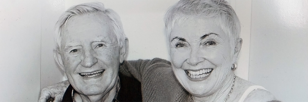 A man and a woman smiling at the camera, the woman, with short hair, has her arm around the man