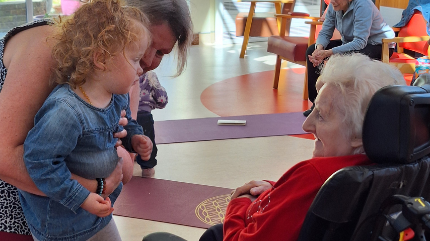 An older lady in a wheelchair looks with a smile at a young child, being held by another lady.