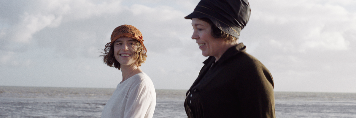 Two women in 1920s dress stand by a body of water, smiling