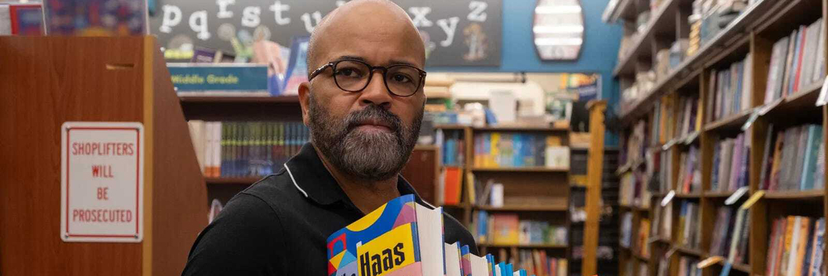 A bald man with glasses is in a bookstore or library holding a stack of books