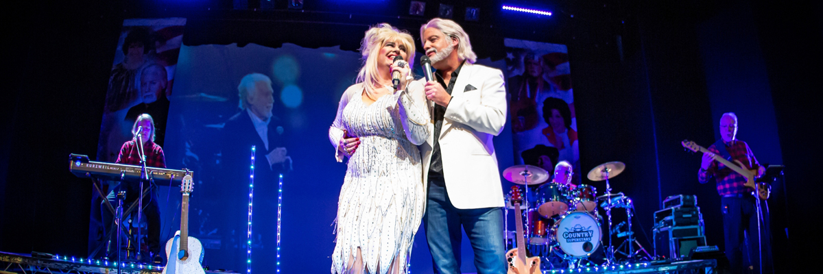 A band is on stage, lit blue. Two people dressed as country singers in white stand with mics to the front of the stage