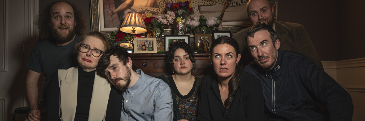 Seven people sit in a line in a dark, old fashioned living room. Some express joy, sadness and irritation.