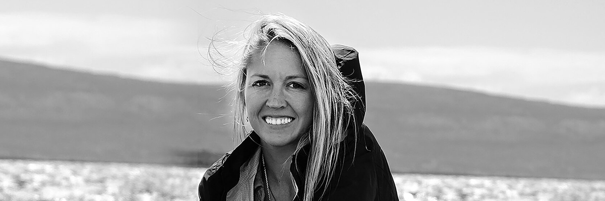 A black and white photo of a blonde lady smiling at the camera. A large lake and mountains are in the background.