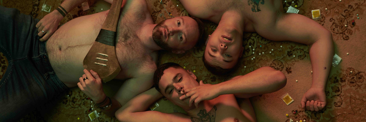 Three men lay face up on a dirty carpet, surrounded by pills. They're all shirtless. One holds a Hurley sliotar, one is smoking.