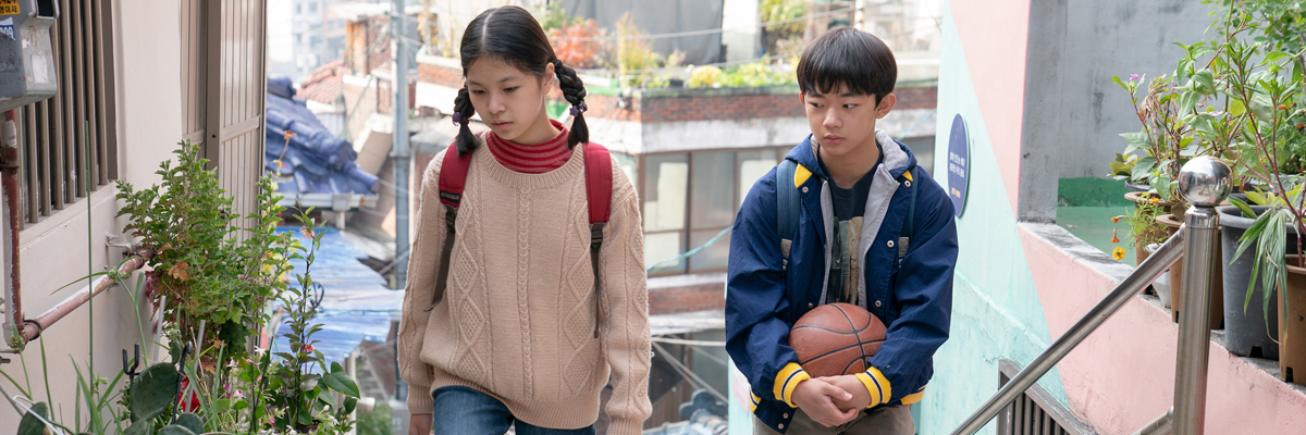 Two young kids walk up a stairway in a residential area. One kid has pigtails, the other kid carries a basketball.