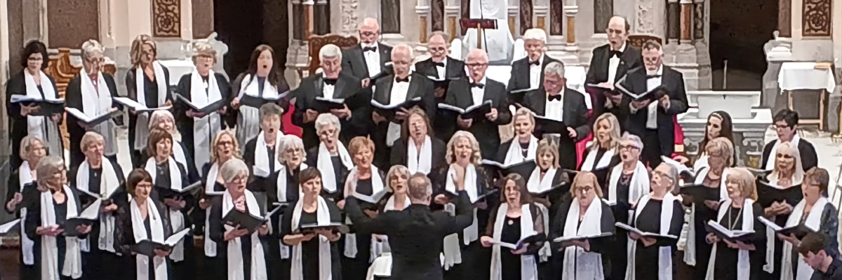 A group of people standing at the front of a church. They are all singing from black bound hymn sheets with a conductor leading in front.