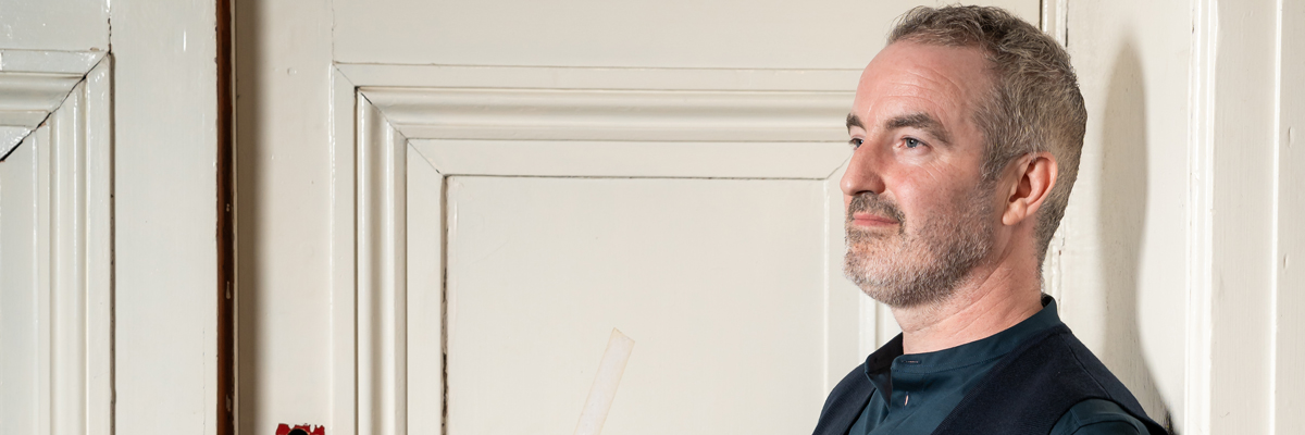 A man with short hair and a beard stares to the left. He leans up against a beige wooden door.