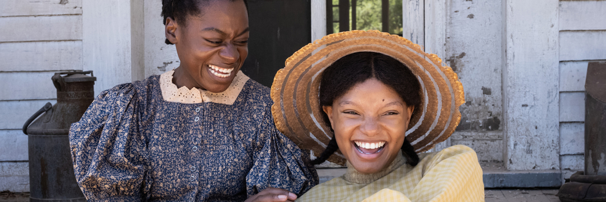 Two women sit on a porch, dressed in 1900's American dresses, laughing together