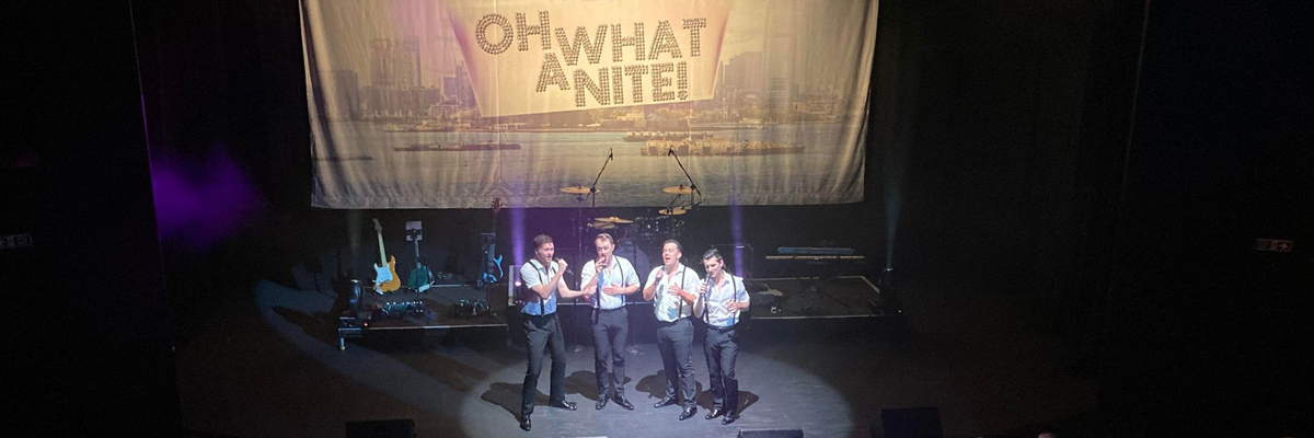 Four men in white shirts, black trousers and suspenders stand on a stand, each singing into microphones. A banner with 'Oh What a Nite! is behind them.