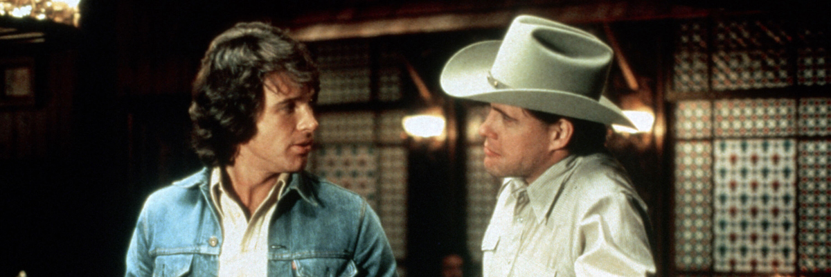 An old looking colour photo of two men looking at each other in a bar. One man wears a beige cowboy hat, the other has shoulder length hair and a denim jacket