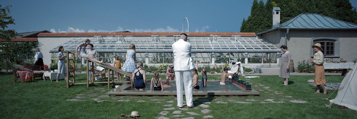 A sunny garden scene, with a greenhouse in the background. People dressed in 1940s clothes and swimming costumes play next and in a small pool with a slide.