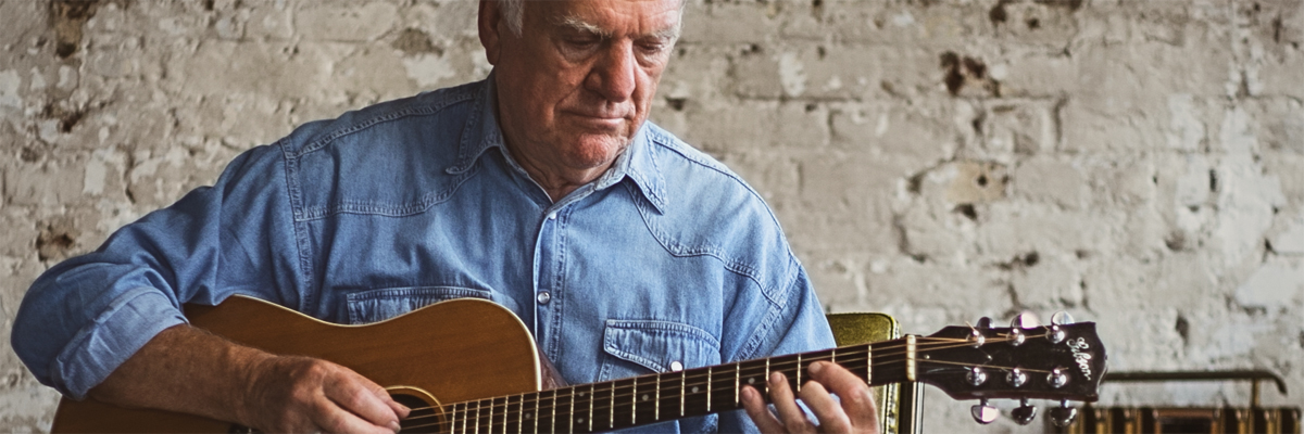 Older grey haired man, wearing a denim shirt, playing an acoustic guitar.